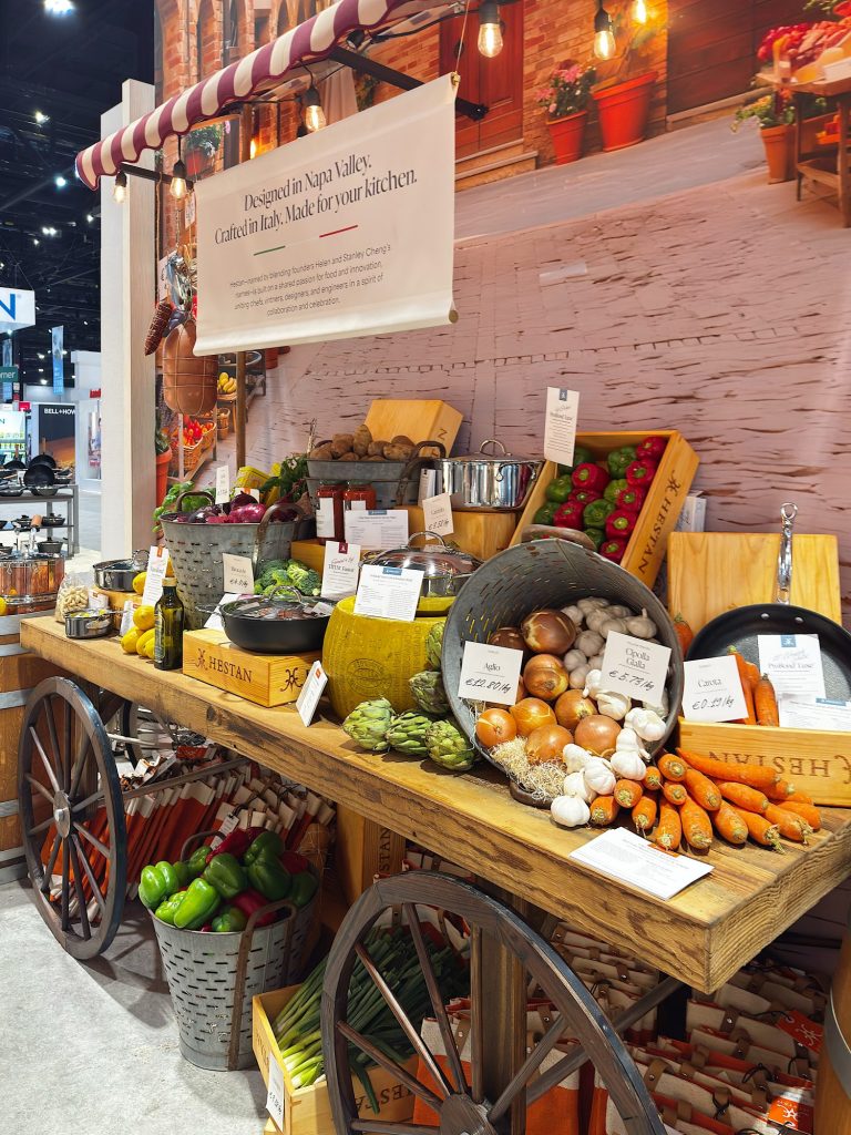 A wooden cart displays various fresh vegetables and herbs in baskets and crates, with labeled signs, at an indoor market or trade show booth.