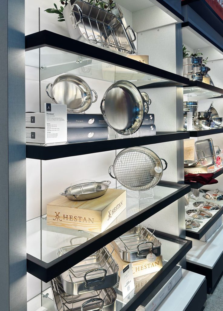 Shelves display stainless steel cookware, strainers, and roasting pans, some labeled Hestan, with product descriptions and wooden boxes.