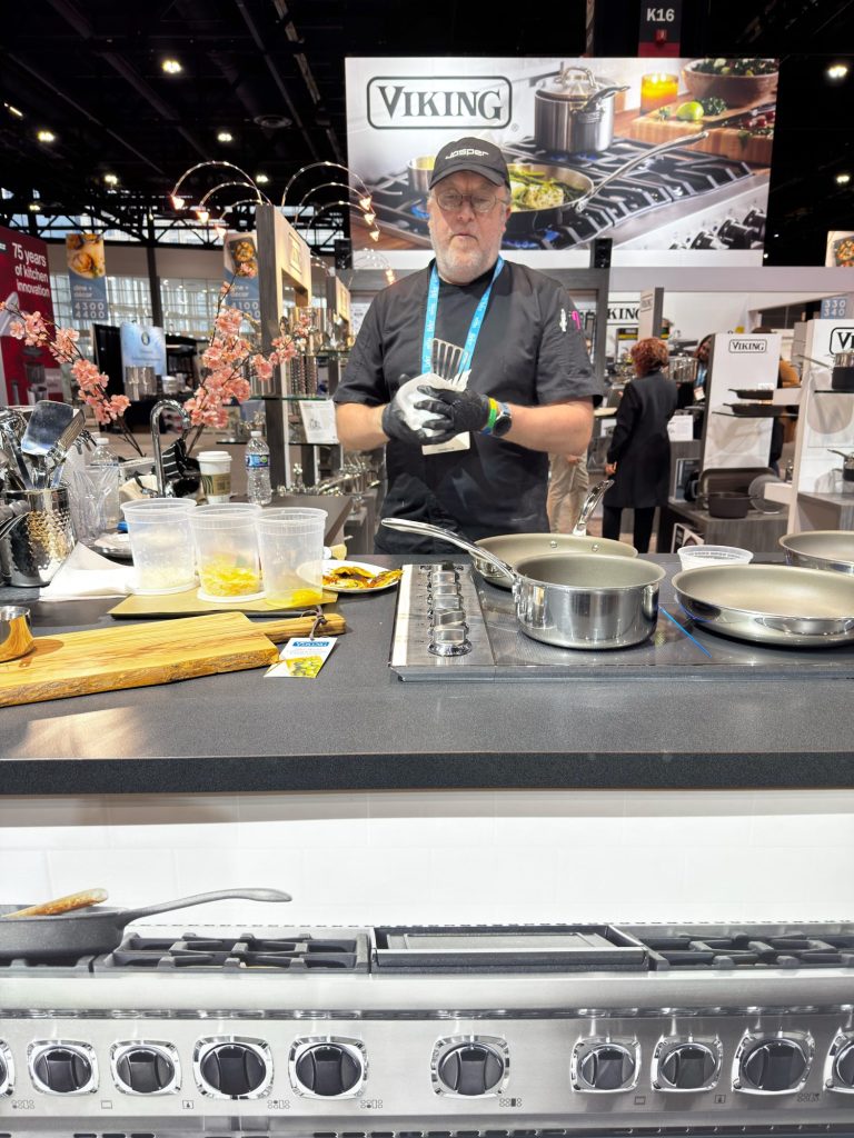A chef wearing a black hat and apron stands behind a kitchen counter with cookware, utensils, and food, at a Viking appliances booth in a convention center.