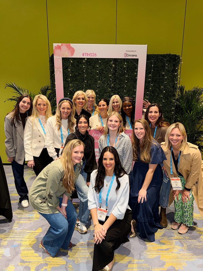 A group of fourteen women pose together indoors in front of a green wall with a pink event frame and badges around their necks.