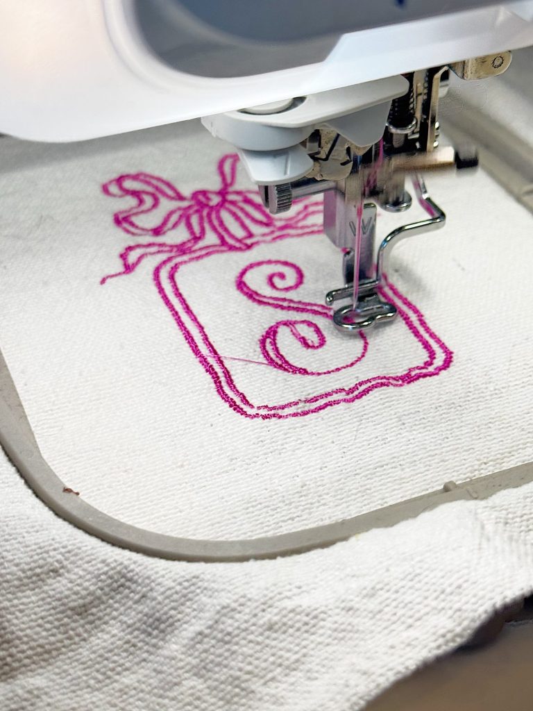 A sewing machine is embroidering a pink design with a letter "E" and a bow onto a piece of white fabric.