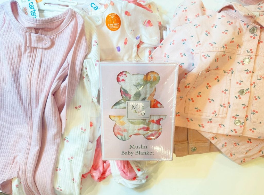 Assorted baby clothes in pink and white patterns are laid out around a boxed muslin baby blanket with a teddy bear design.