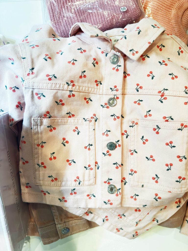 A light pink baby jacket with small red cherry prints and silver buttons, displayed with matching clothing items.