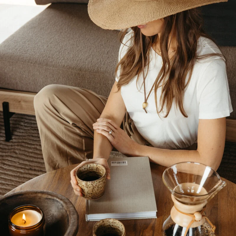 A woman in a wide-brimmed hat and white shirt sits on the floor by a table with coffee, a candle, and a closed notebook.
