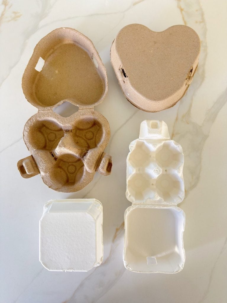 Four small cartons, two made of brown cardboard and two of white foam, are open to show their interiors on a marble surface.