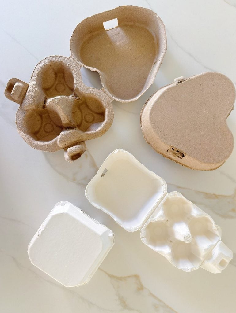 Four small, heart-shaped and rectangular egg cartons, two made of brown cardboard and two made of white material, are displayed open on a white marble surface.