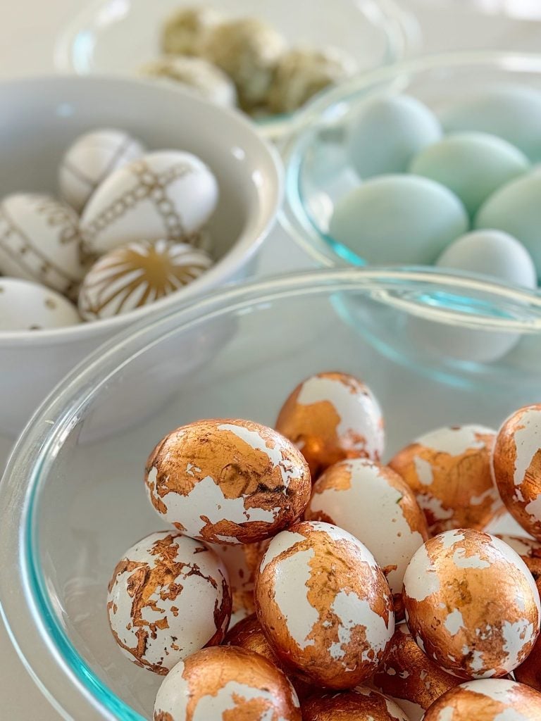 Four glass bowls filled with decorated Easter eggs, including eggs with gold foil, light blue eggs, and white eggs with brown geometric designs.
