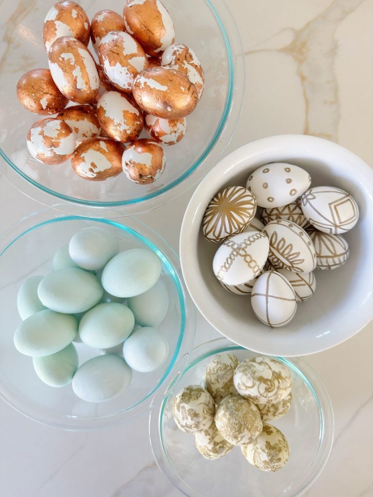 Four glass bowls filled with decorated eggs: copper-leaf eggs, blue eggs, eggs with gold geometric designs, and eggs with gold marbled patterns on a marble surface.