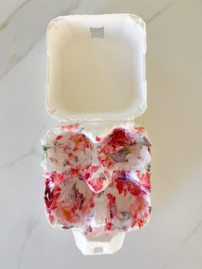 An open egg carton contains melted and swirled red, white, and pink crayon wax in the cups.