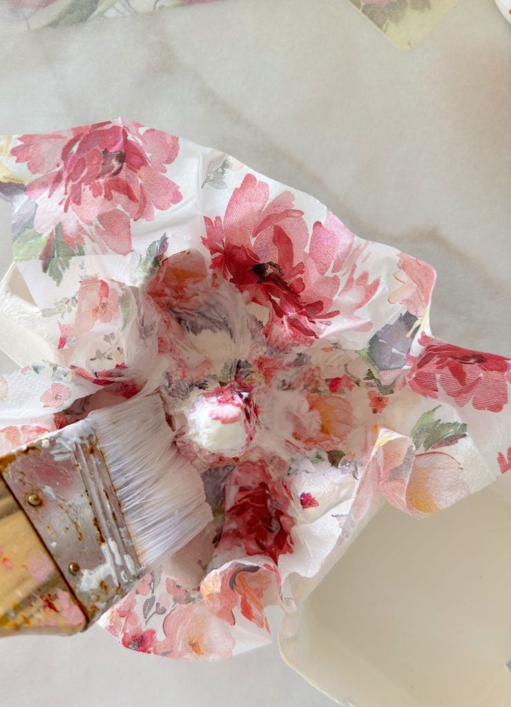 A hand uses a paintbrush to apply glue or paste to floral-patterned fabric draped over an upside-down bowl, likely for a craft project.