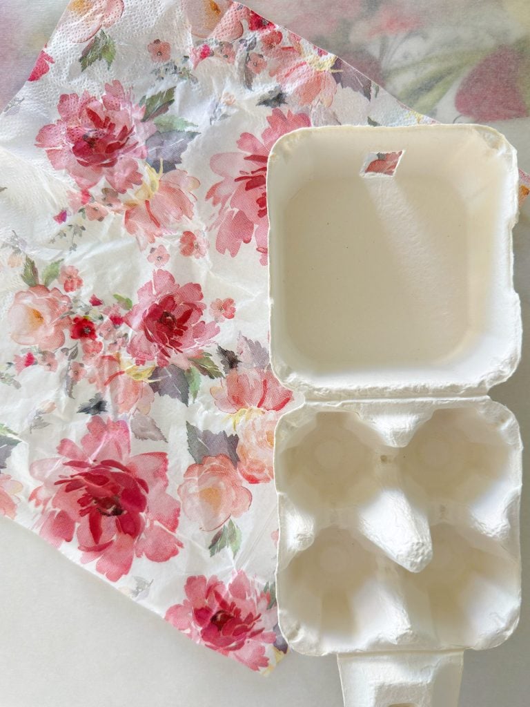 An empty white egg carton is open and placed next to a floral-patterned napkin with red and pink flowers on a light surface.