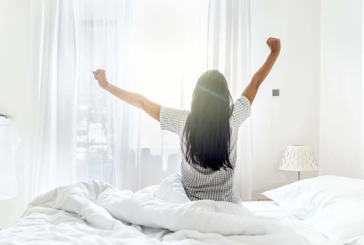 A person with long hair sits on a bed, facing a bright window, stretching their arms in the morning light.