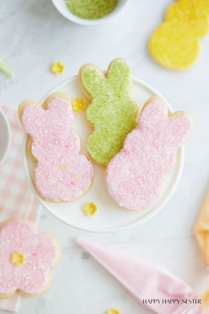 Three bunny-shaped sugar cookies, two frosted with pink sprinkles and one with green sprinkles, are displayed on a white cake stand.