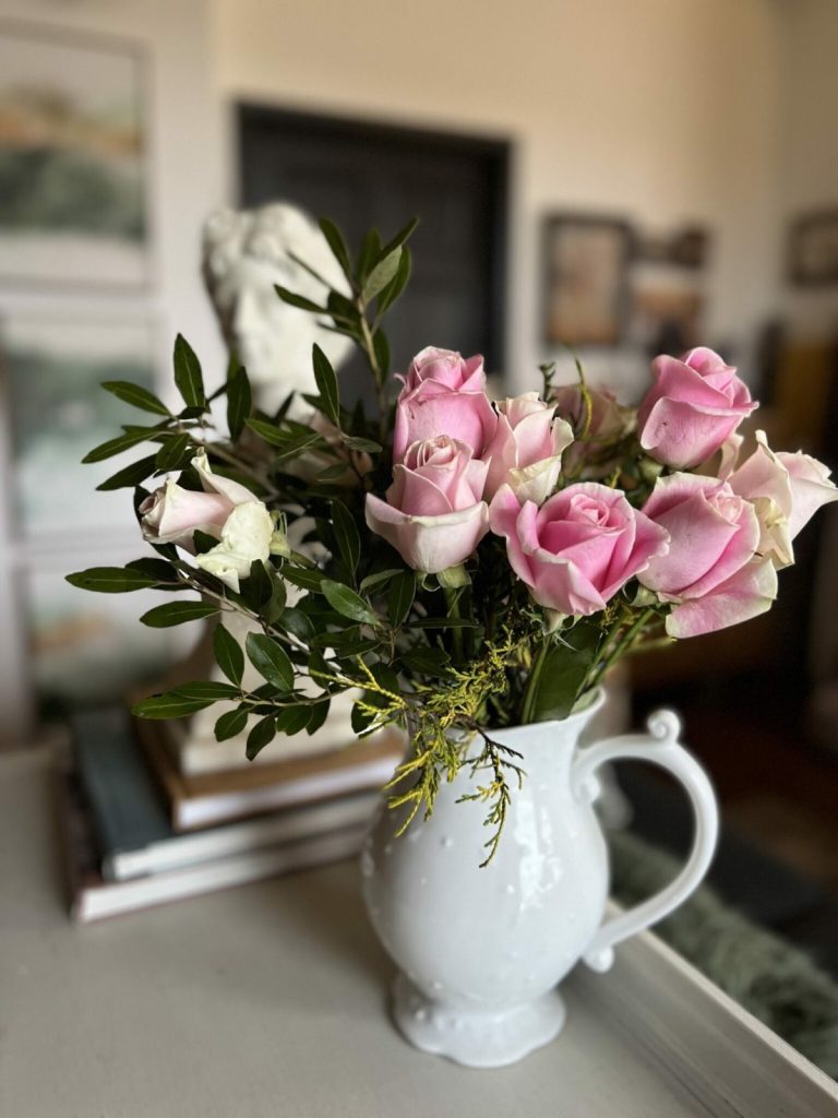 A white ceramic pitcher filled with pink and white roses and green foliage sits on a table beside a stack of books in a softly lit room.