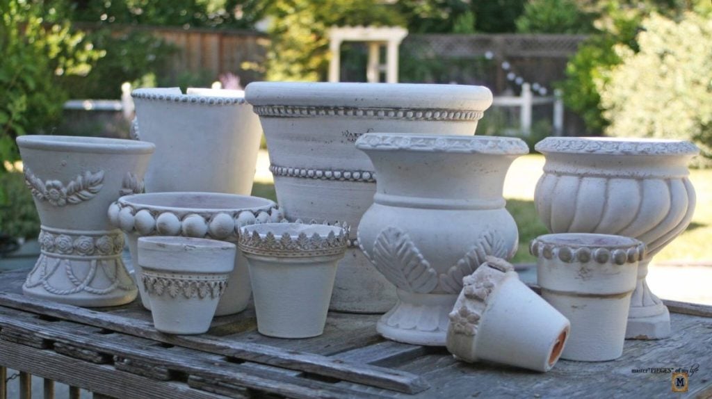 A group of ornate, white ceramic flower pots of varying sizes and designs are arranged on a wooden surface outdoors.