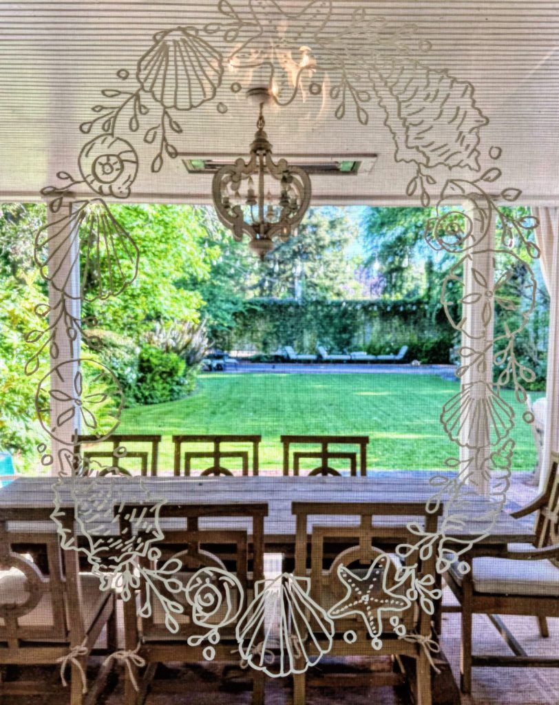 A dining area with wooden chairs and table overlooks a backyard with green grass, pool, and lounge chairs; white seashell, window wreaths, and marine designs frame the image.