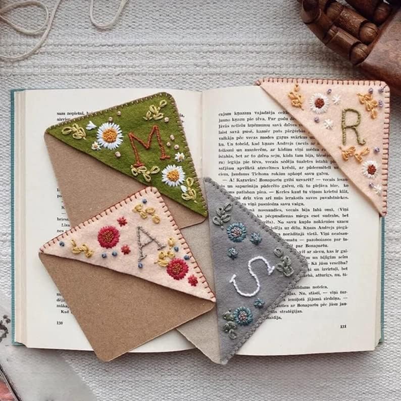 Four embroidered felt corner bookmarks, each with floral designs and a different letter (M, A, S, R), rest on the pages of an open book.