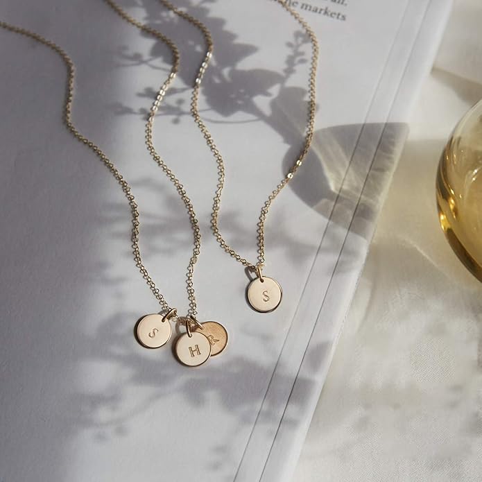 Three gold necklaces with round initial pendants&mdash;S, H, and S&mdash;are arranged on an open book, with shadows and a glass object nearby.