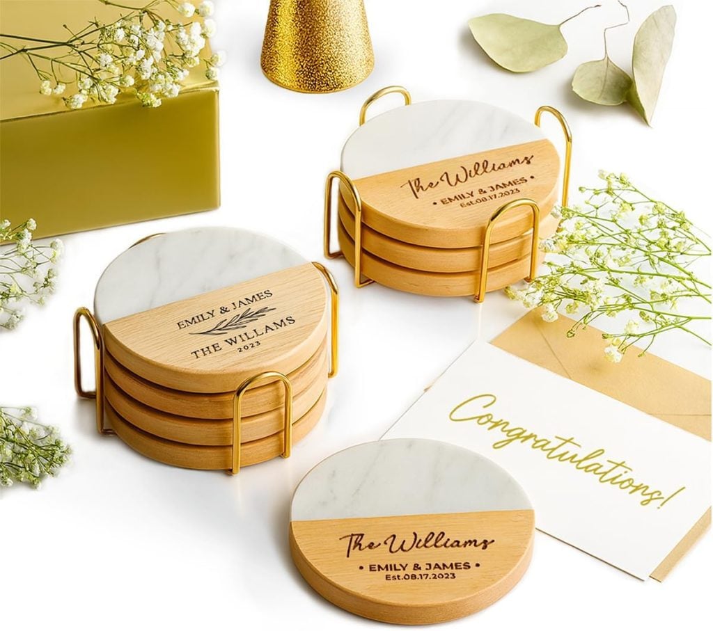 Two sets of round marble and wood coasters with personalized engravings&mdash;perfect for adding a name for your home&mdash;rest in gold holders beside baby's breath, a gold envelope, and a "Congratulations!" card.
