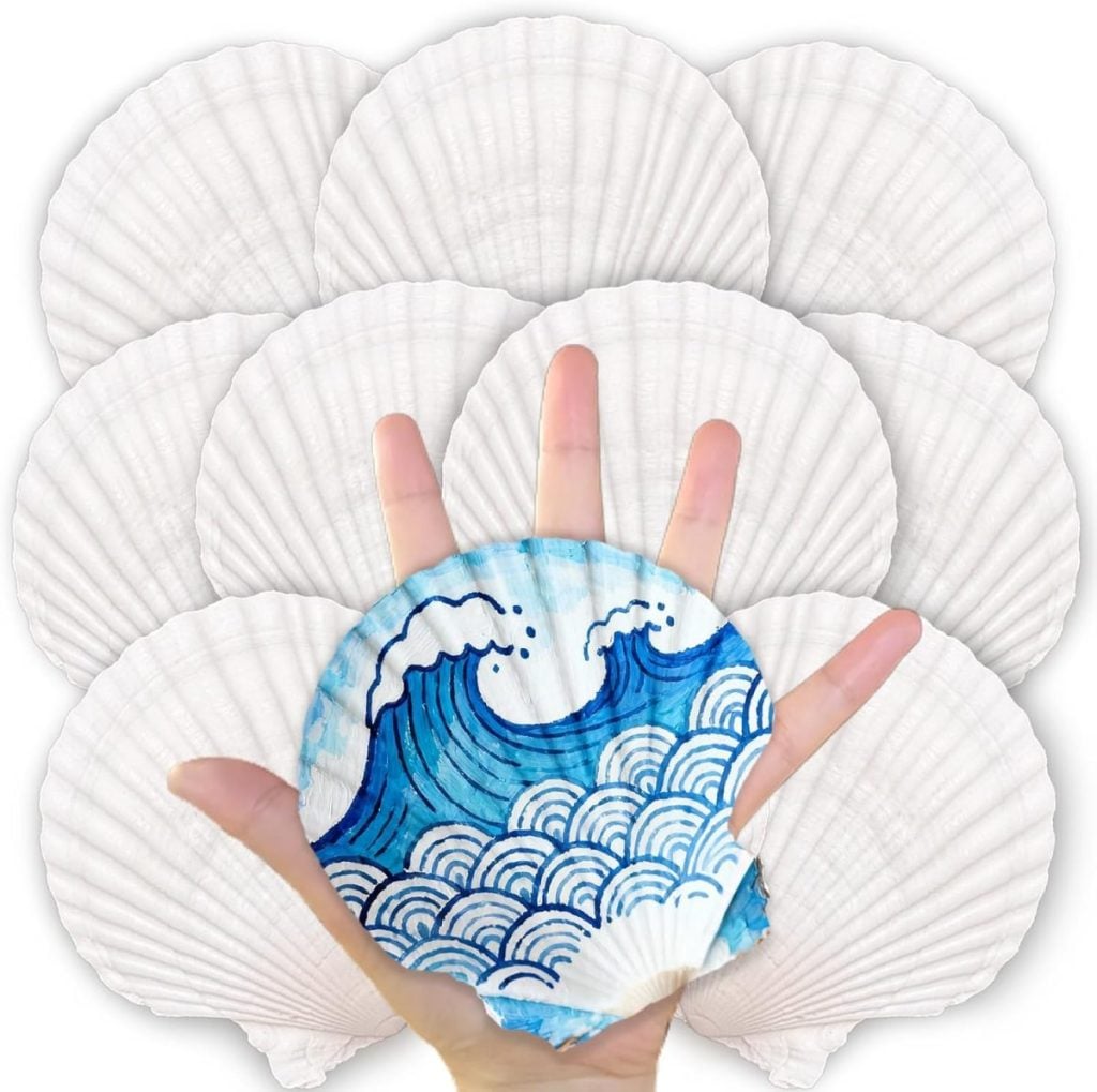 A hand holds a handpainted shell featuring a blue ocean wave design, with several plain white seashells fanned out in the background.
