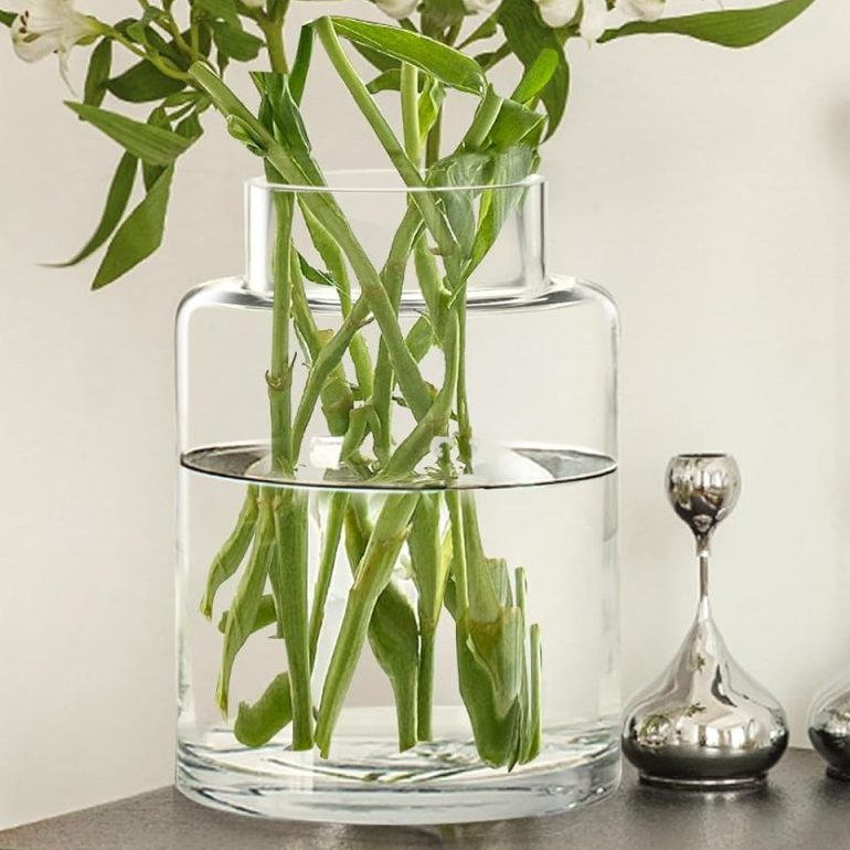 A clear glass vase with white flowers and green stems sits on a dark table next to two silver decorative objects, creating a serene Airbnb vibe in Waco with a bed visible in the background.