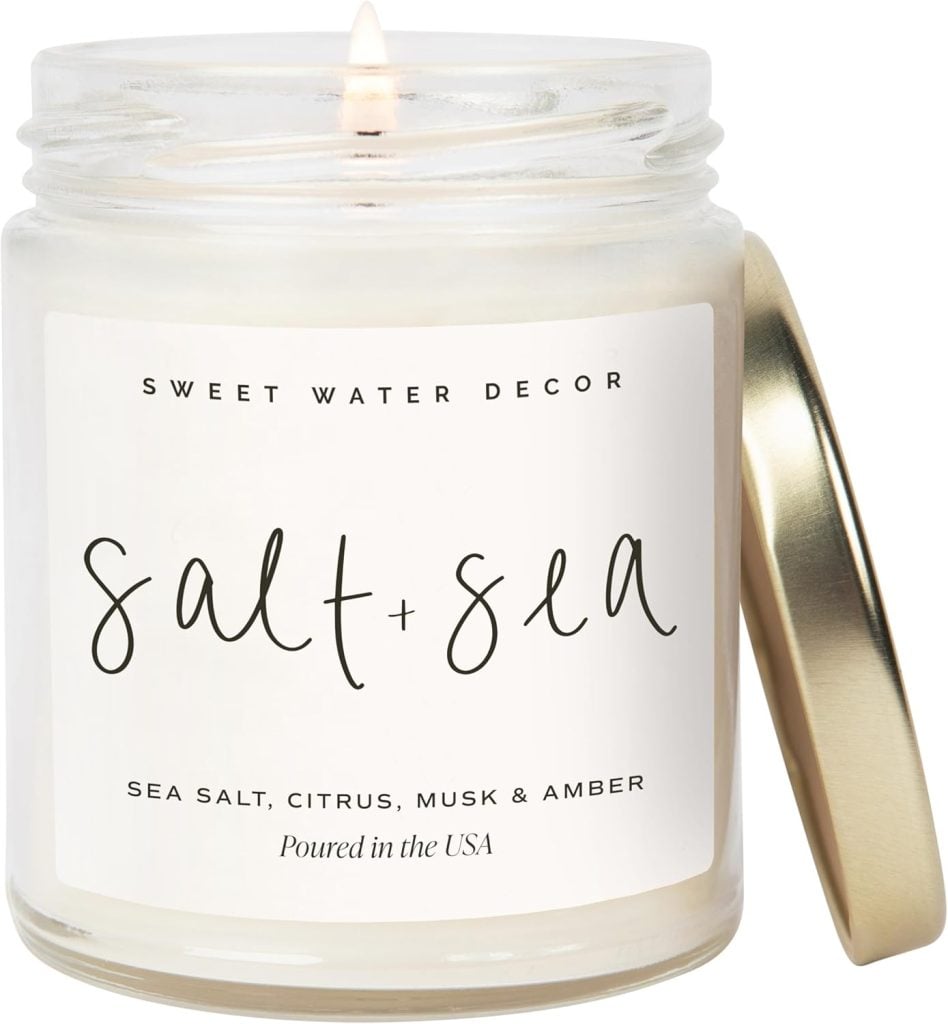 A lit white candle in a glass jar labeled "salt + sea," featuring scents of sea salt, citrus, musk, and amber. The perfect touch for Mother's Day Gifts Amazon, the jar has a gold lid resting beside it.
