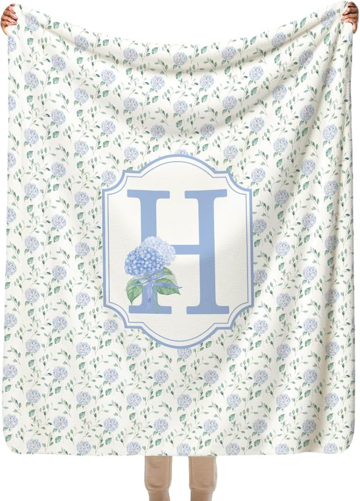 A person holds a blanket featuring a large blue letter "H" and a hydrangea flower design, with a pattern of blue hydrangeas and green leaves in the background.