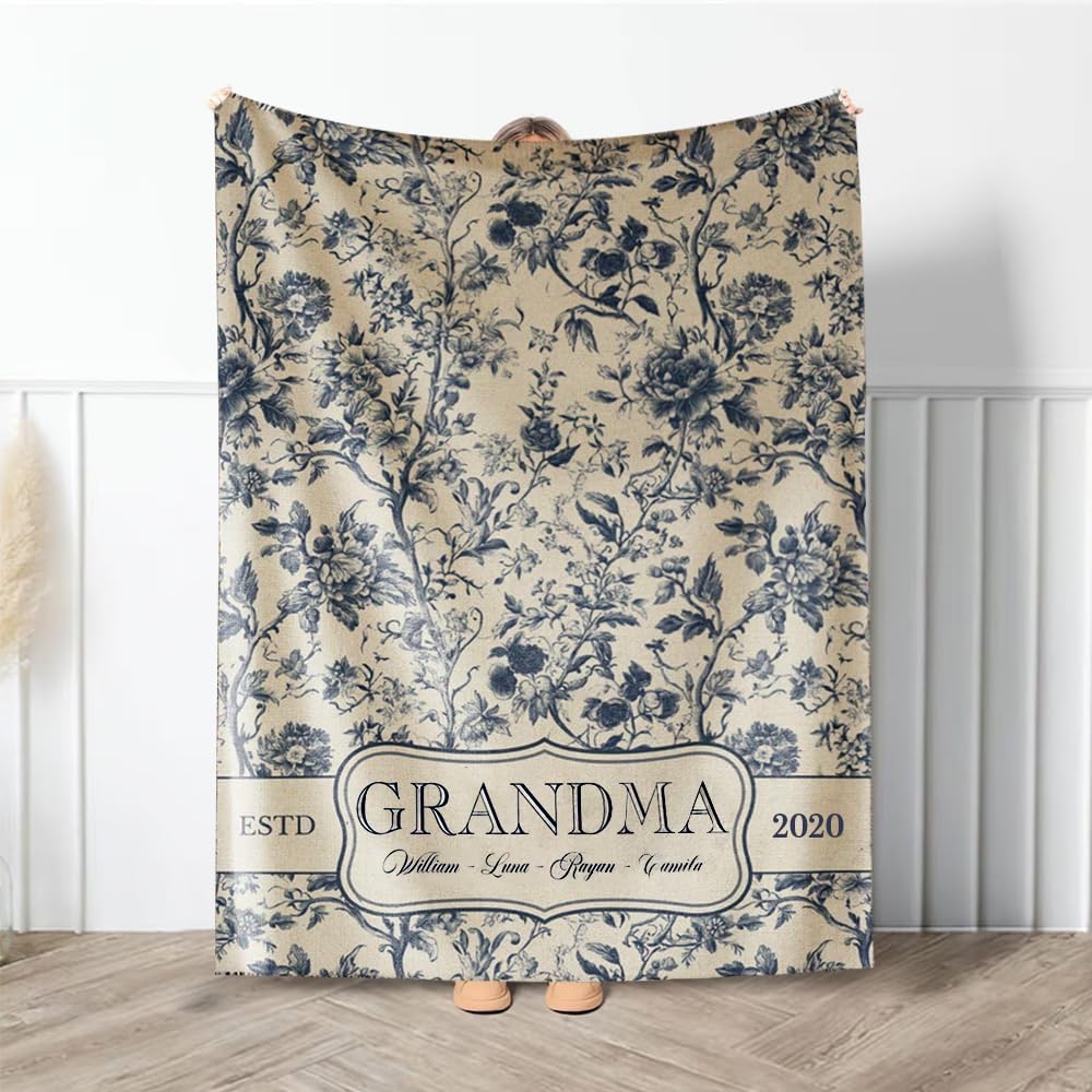 A person holds up a beige and blue floral-patterned blanket with the text "GRANDMA," "ESTD 2020," and four names printed along the bottom.