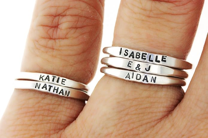 Close-up of four silver rings on fingers, each ring engraved with different names: Katie, Nathan, Isabelle, E&J, and Aidan.