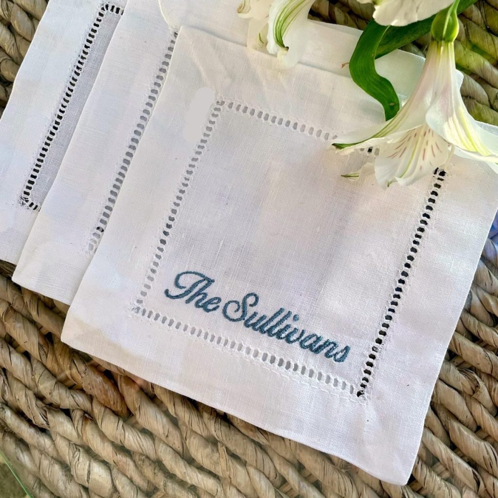 White embroidered napkin with the text "The Sullivans" placed on a woven surface, partially covered by white flowers.
