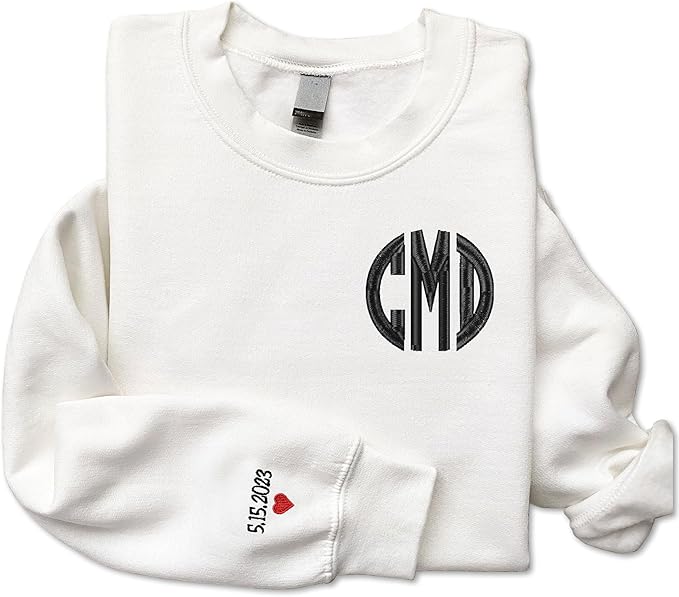 White sweatshirt with black "CMD" monogram on the chest; left sleeve features the date "5.15.2023" and a small red heart near the cuff.