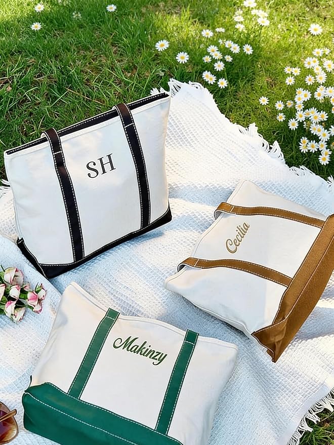 Three canvas tote bags with different colored handles and personalized names or initials sit on a white blanket on grass with daisies.