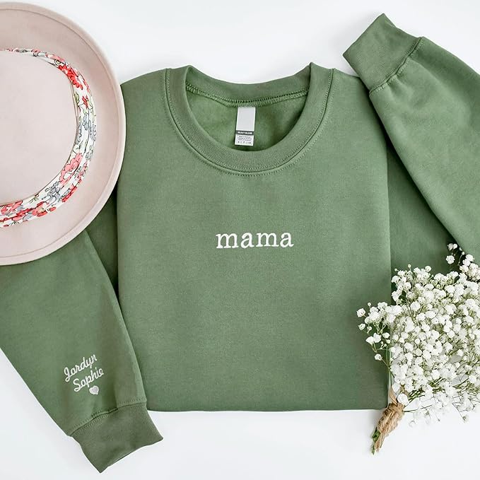 A green sweatshirt with "mama" printed in white on the front, next to a pink hat with floral lining and a small bouquet of white flowers. The sleeve is embroidered with names.