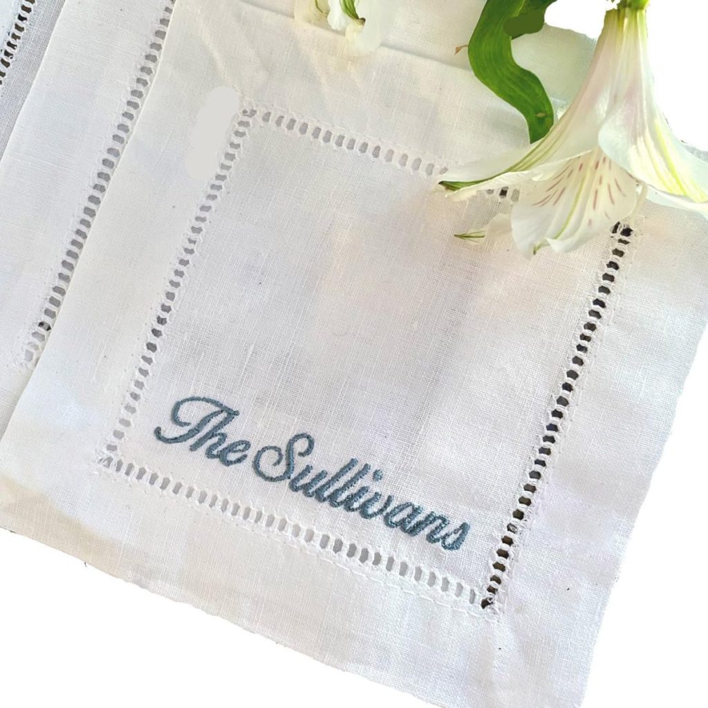 A white linen napkin with "The Sullivans" embroidered in blue script, bordered by a decorative hemstitch, with white flowers partially visible at the top right.