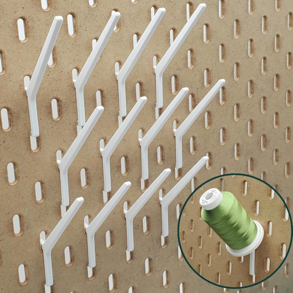 White plastic pegboard hooks are mounted on a brown perforated board, with one hook&mdash;perhaps prepping supplies for an east coast trip&mdash;holding a spool of light green thread in the lower right corner.