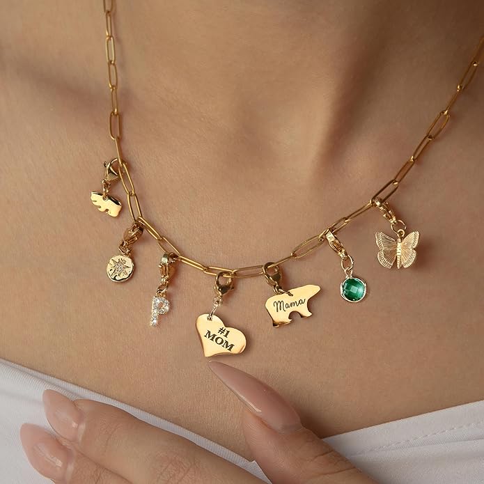 A close-up of a gold chain necklace with various charms, including hearts, a butterfly, a green gem, and the words &ldquo;#1 MOM&rdquo; and &ldquo;Mama,&rdquo; worn around a woman&rsquo;s neck.