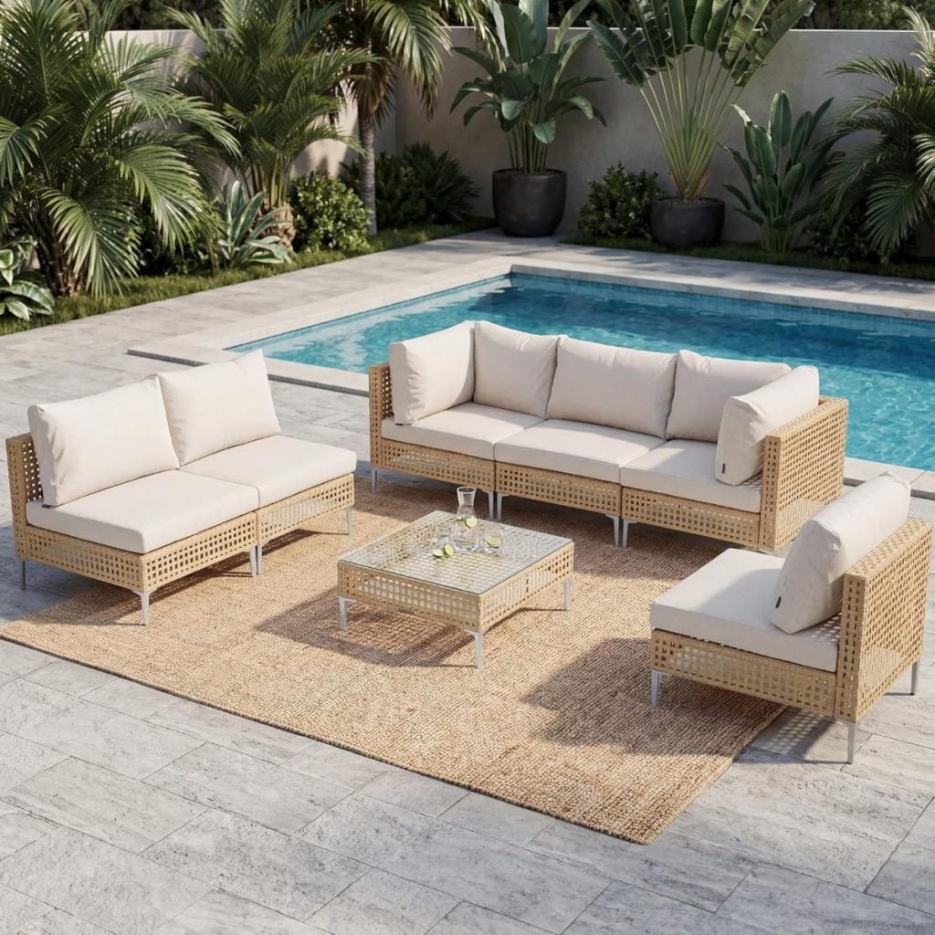 Outdoor patio set with cream cushions on wicker sofas and a glass-top coffee table, arranged on a woven rug beside a swimming pool and surrounded by tropical plants.