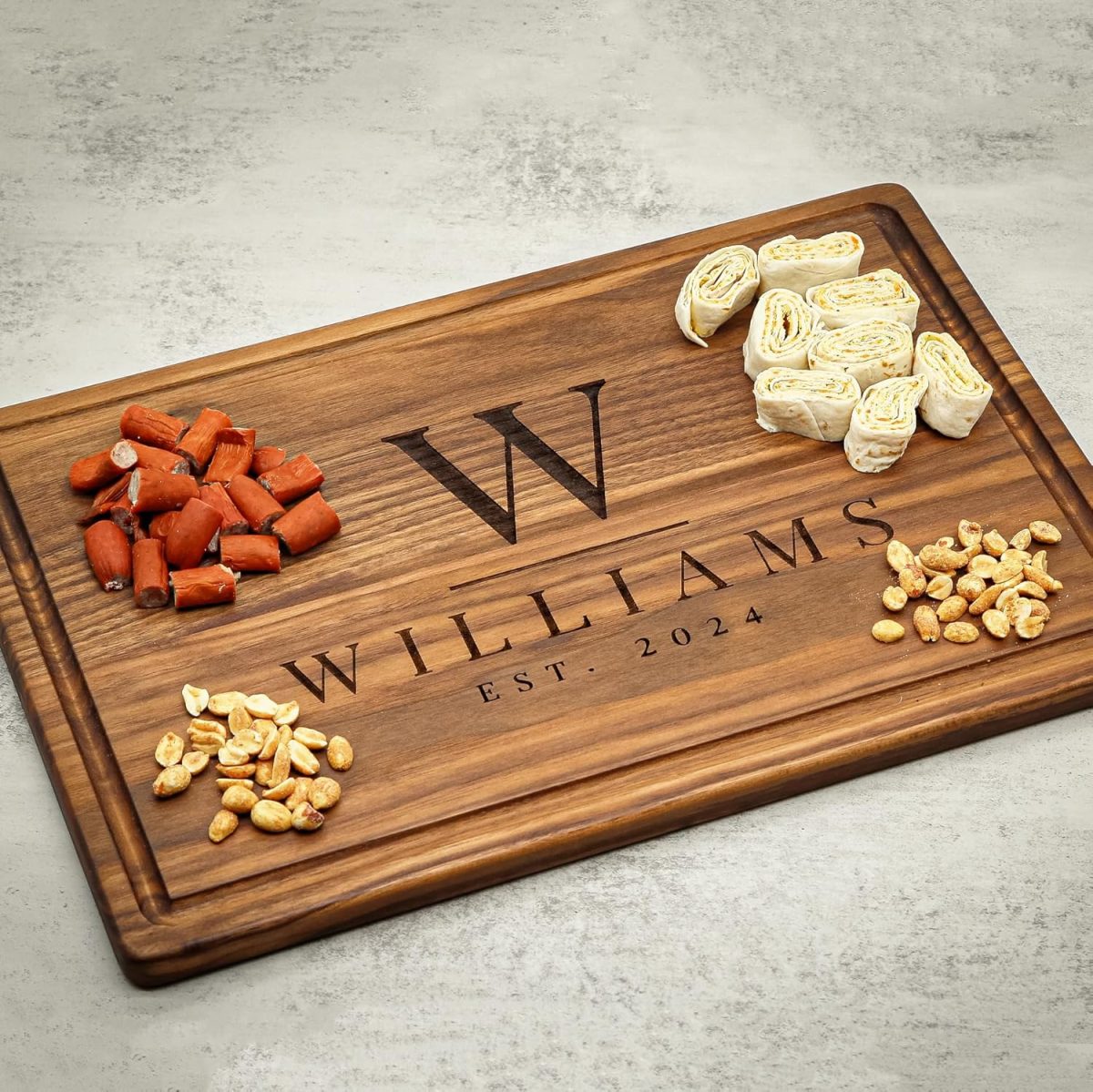 A personalized wooden cutting board labeled "Williams Est. 2024" with diced sausage, rolled cheese slices, and peanuts arranged on top.