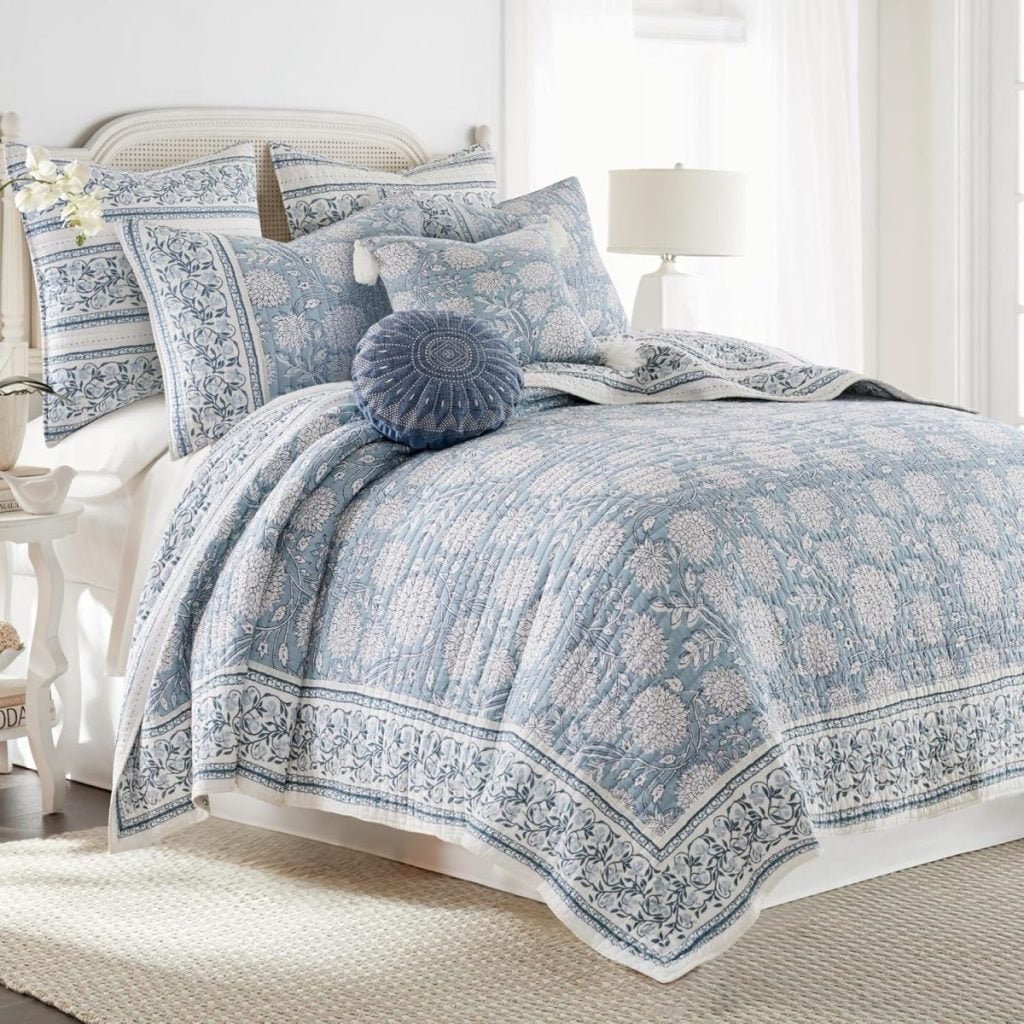 A neatly made bed with a blue and white floral quilt, multiple pillows, a round blue cushion, and a white nightstand with a lamp in a bright, airy bedroom&mdash;perfect inspiration for Mother&rsquo;s Day gifts on Amazon.