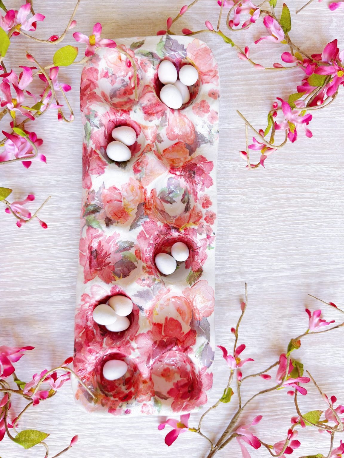 A floral-patterned egg carton holds small white eggs, surrounded by pink and green artificial flower branches on a light wooden surface.