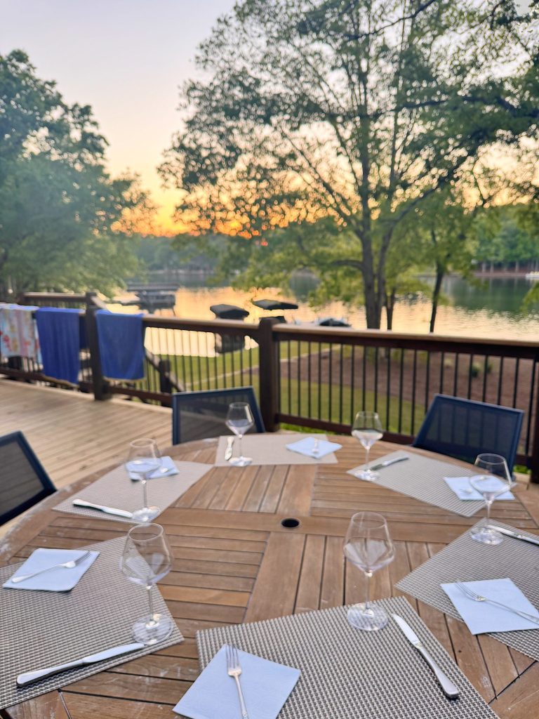 A round wooden outdoor table set with placemats, napkins, cutlery, and wine glasses on a deck overlooking a lake at sunset.