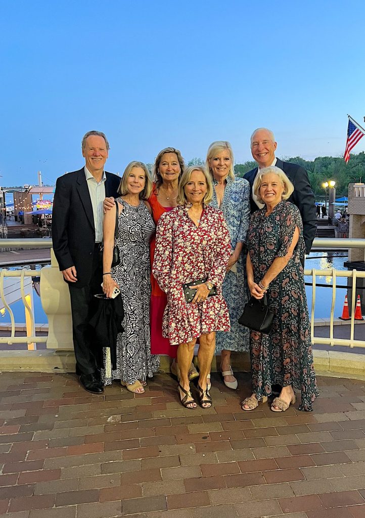 A group of seven adults, four women and three men, stand together smiling outdoors by a waterfront in the evening. An American flag is visible in the background.