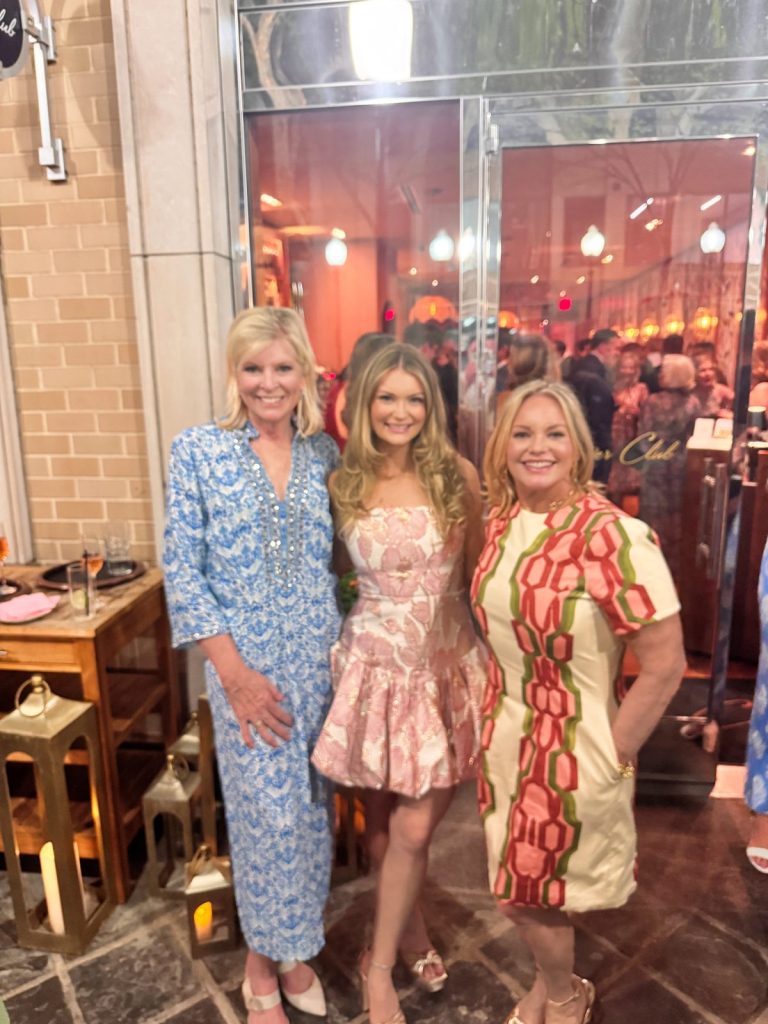 Three women stand and smile at an indoor event, wearing patterned dresses. There are reflections and people visible through the glass doors behind them.
