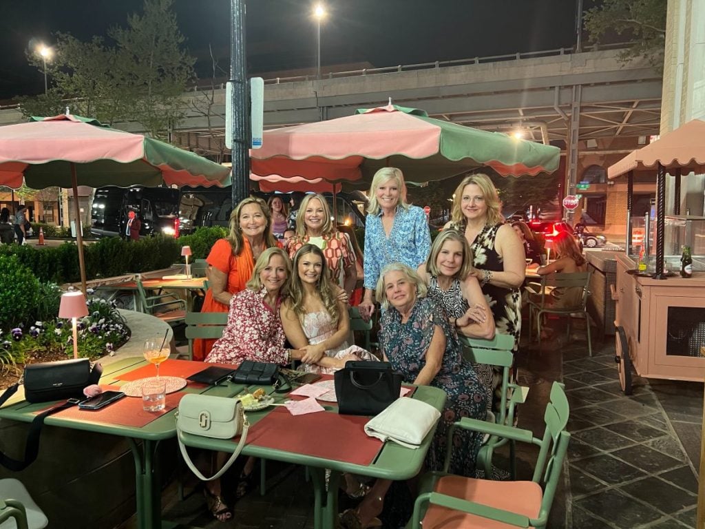 A group of nine women sit and stand around a table at an outdoor restaurant at night, smiling at the camera. Some purses and drinks are visible on the table.