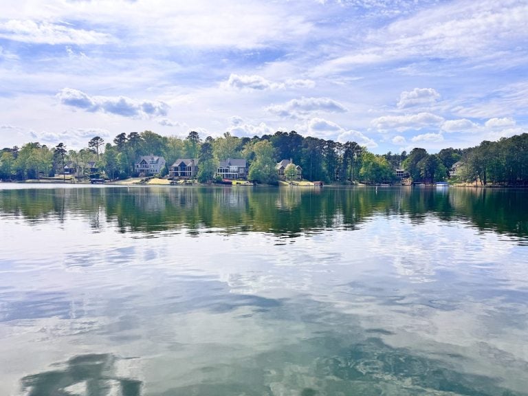 Houses and trees line the far shore of a calm lake under a partly cloudy sky, with reflections visible in the water.