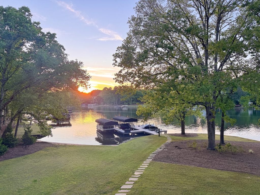 A lakeside scene at sunset with boats docked at a pier, large trees, calm water, and a stone path leading to the shore.