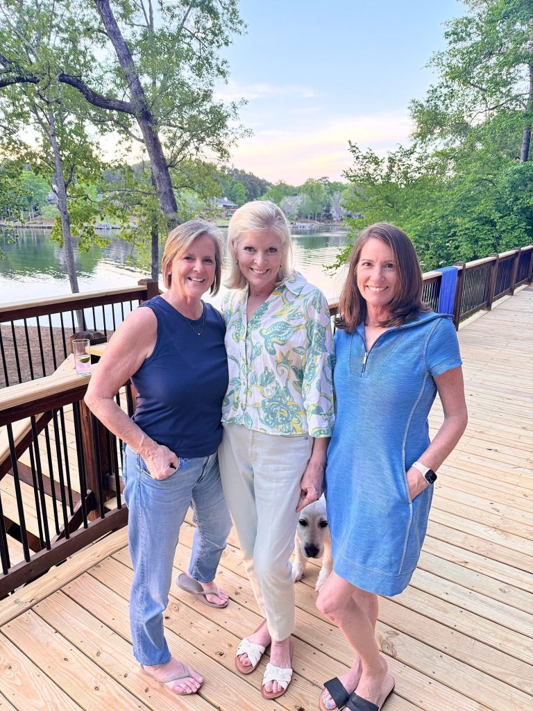 Three women stand and smile on a wooden deck by a lake, with trees in the background and a dog partially visible between them.
