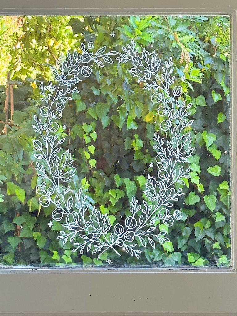 A window with a white, hand-drawn wreath design on the glass, with green ivy and foliage visible outside.