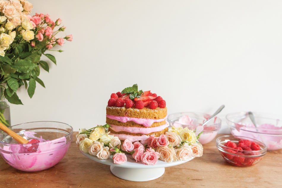 A layered cake with pink frosting and berries sits on a white cake stand surrounded by roses, with bowls of frosting and raspberries nearby on a wooden table.