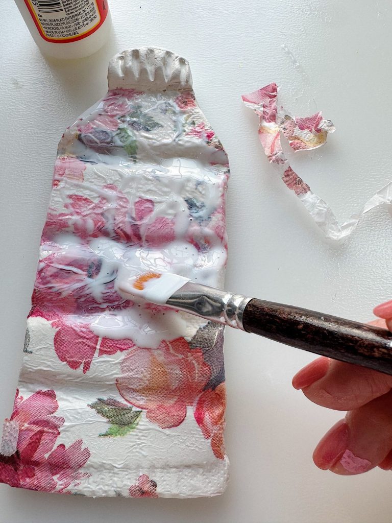 A hand applies glue to a floral-patterned, flattened plastic bottle with a paintbrush; a bottle of glue and torn patterned paper are nearby.